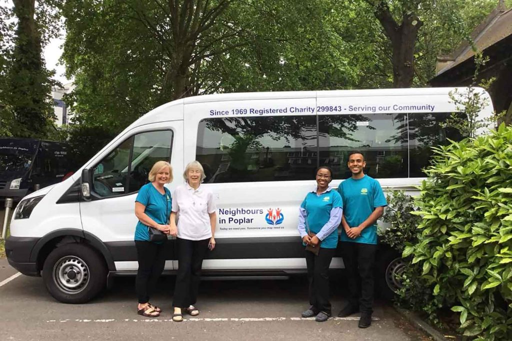 Sister Christine Frost MBE stood infront of the white Neighbours in Poplar minibus with three other members of the Neighbours in Poplar team all smiling.