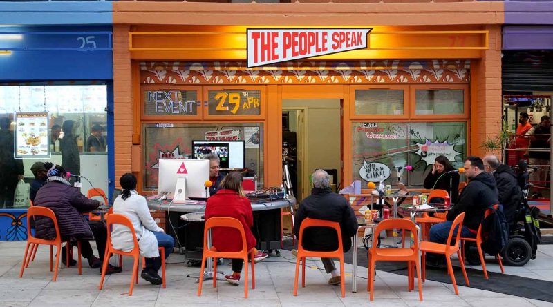 A group of people sat around round tables outside the orange The People Speak space on Aberfeldy Street