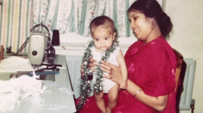 Bangladeshi seamstress Anwara Begum sits at table with sewing machine, holding her baby Asma Begum, East London